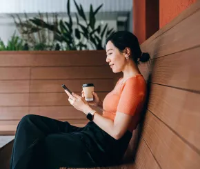 A young professional lady looks at her phone while having a coffee on an outdoor bench